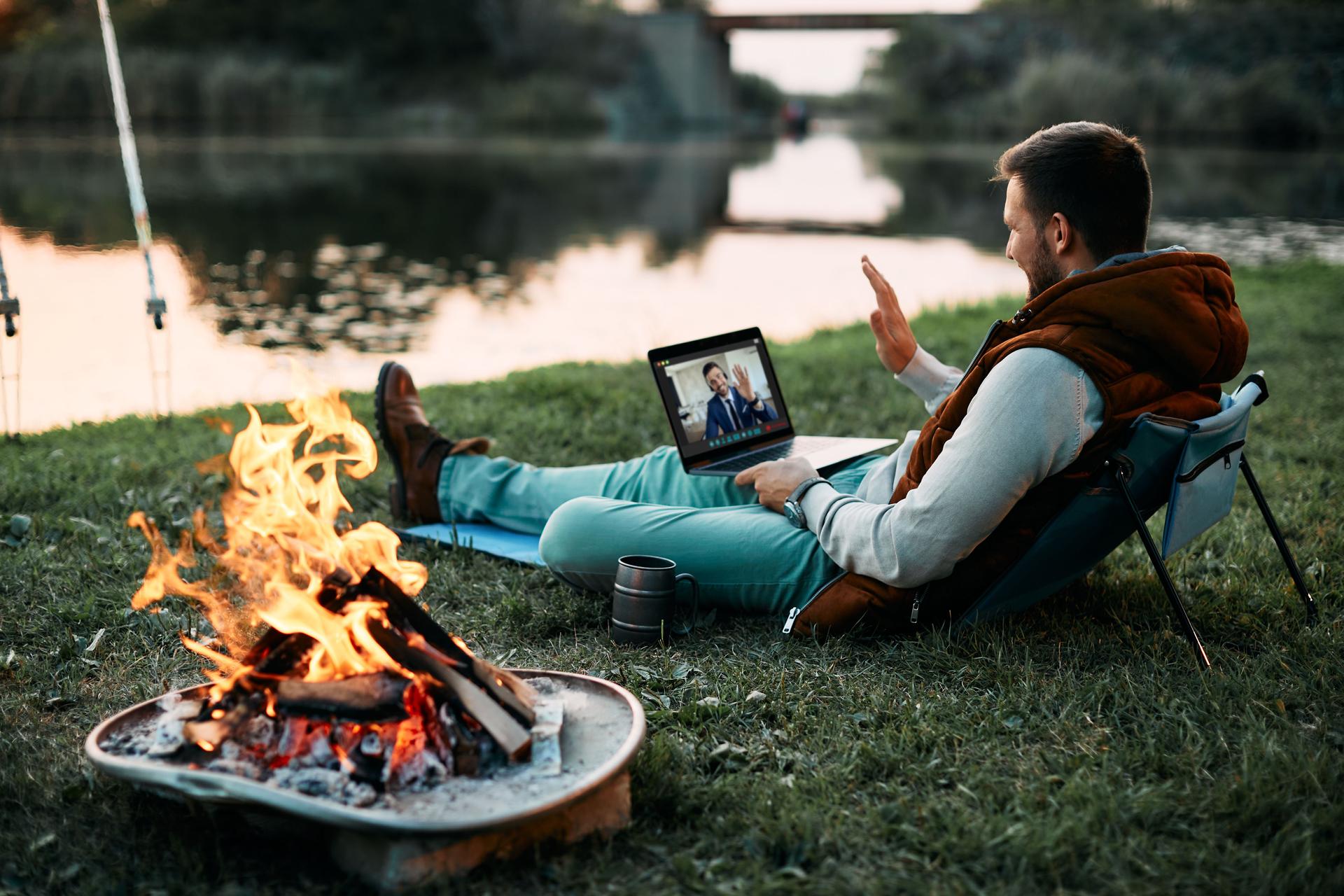 Happy man having video call with business co-worker while relaxing by campfire and fishing in nature.